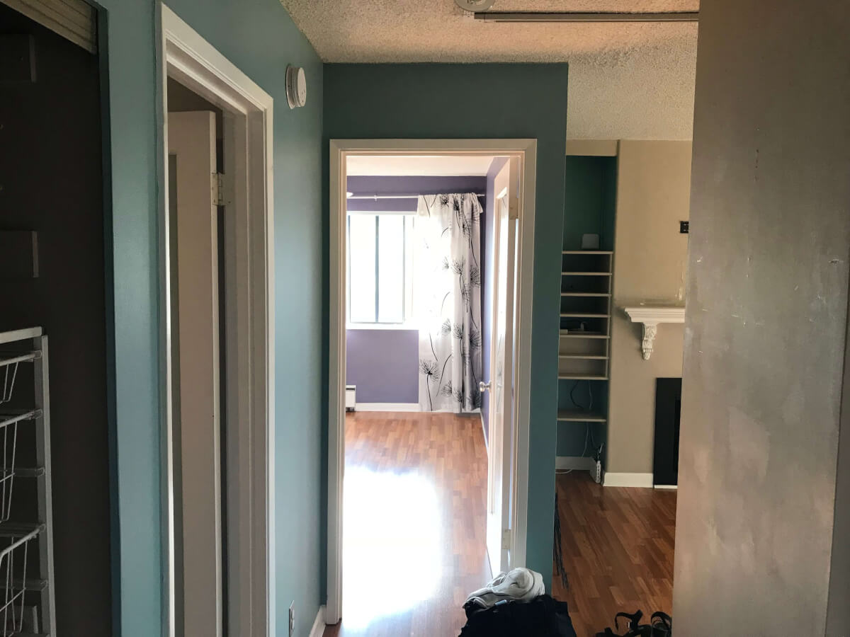 A view from my front door looking through entranceway into the bedroom. Light blue walls. The walls in the bedroom are purple and the flooring throughout is dark brown laminate.