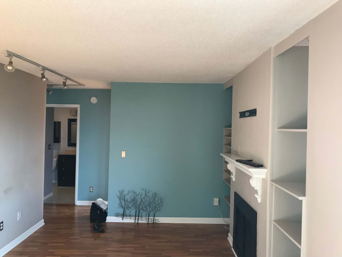 Photo of my lounge area with light blue walls. A black fireplace with a white ceramic mantlepiece above. Above the mantle is an empty black TV bracket. Either side of the fireplace are recessed shelves. The ceiling is off-white and the floor is dark brown laminate.