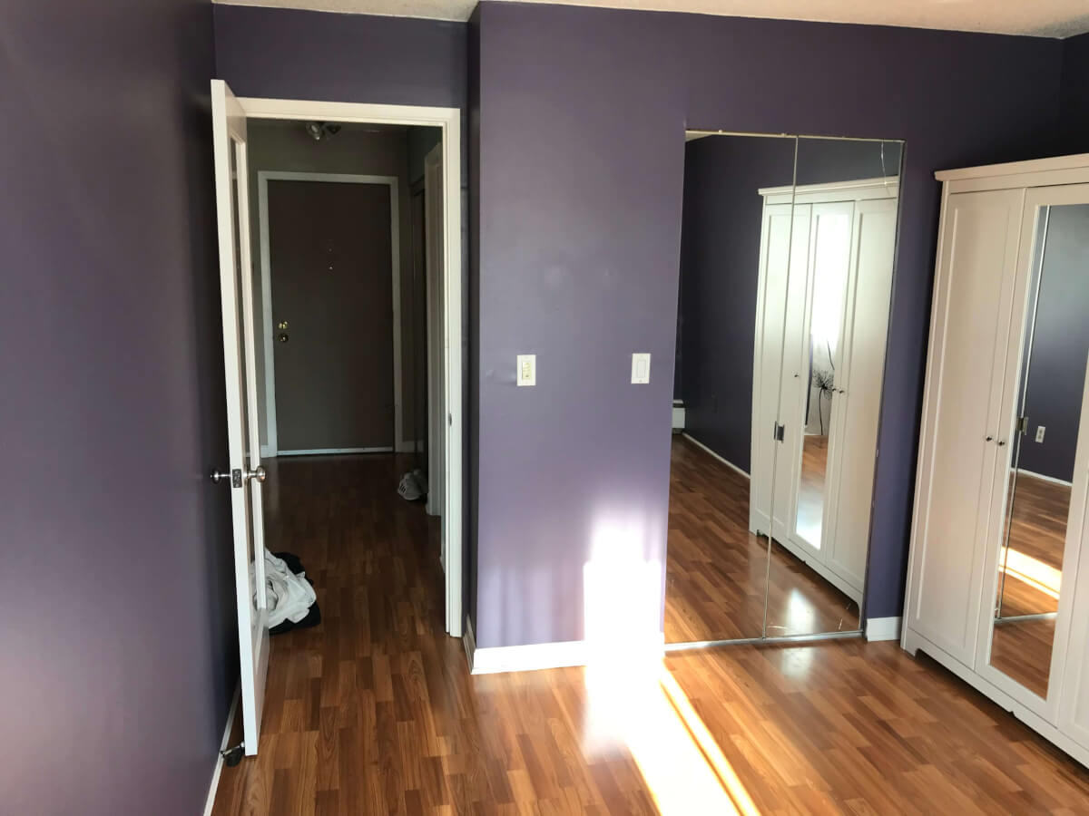 A view from my bedroom looking back towards the entranceway. The walls are purple. The closet bi-fold doors are mirrored. The flooring is dark brown laminate,