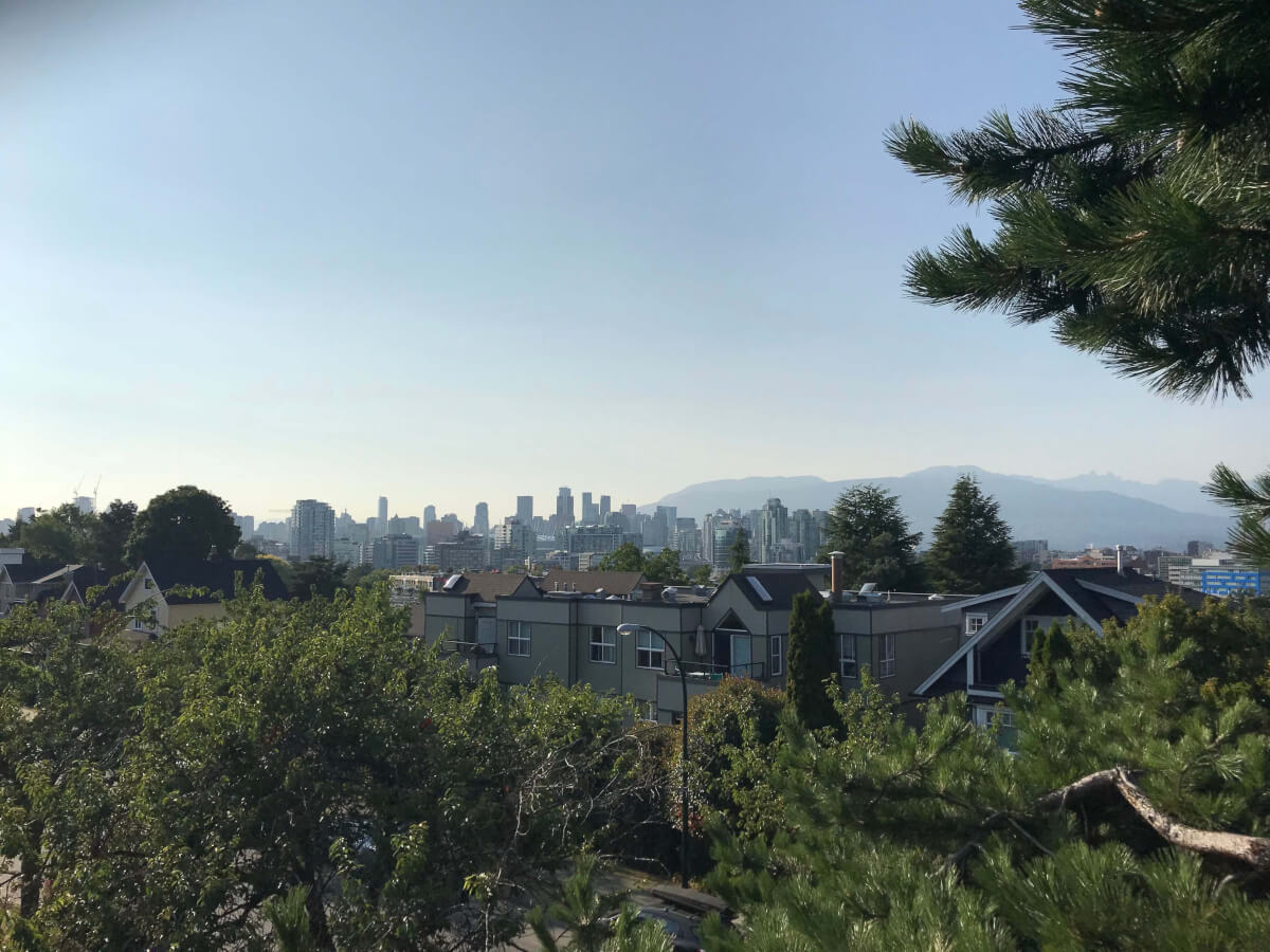 A photo from my balcony of downtown Vancouver with its high-rises as well as the North Vancouver mountains and a brilliant blue sky. In the foreground are lots of trees and some low-rise apartments.